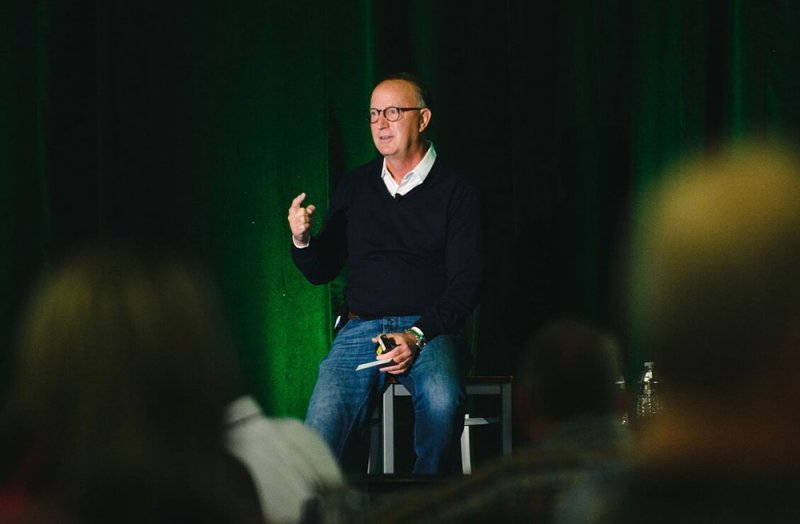 man sitting on stage presenting to a large crowd