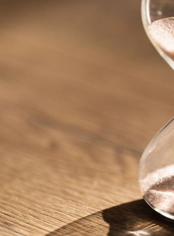 Image of an hourglass sitting on a wooden table; the sand is currently poured halfway, indicating half the time is gone.