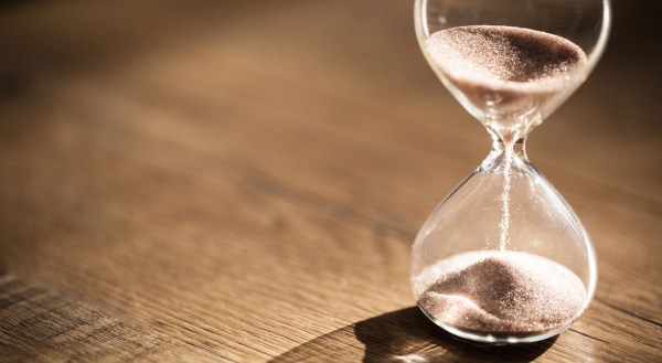 Image of an hourglass sitting on a wooden table; the sand is currently poured halfway, indicating half the time is gone.