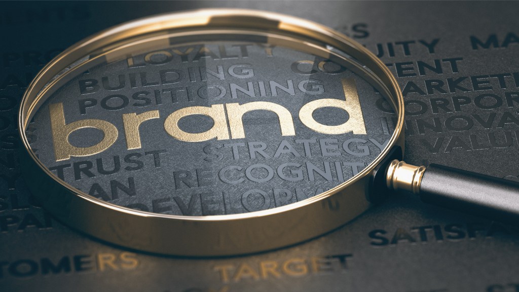 A word cloud sits on a black background. The word “brand” is in gold and is visible under a magnifying glass with a gold handle.